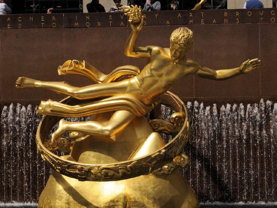 Photo of a golden statue. It is of a man who looks like he is flying and holding fire. It is in Rockerfeller Center in New York City.