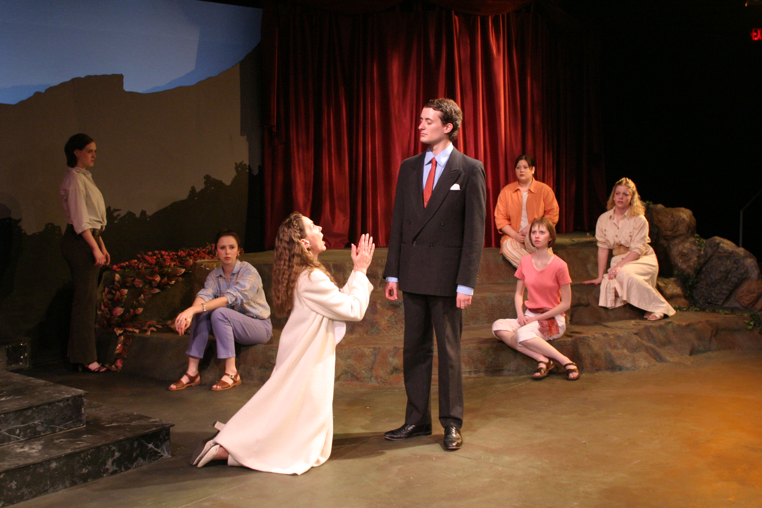 Stage scene with actors; a man in a suit stands while a woman kneels before him. Other actors are seated around them on steps.
