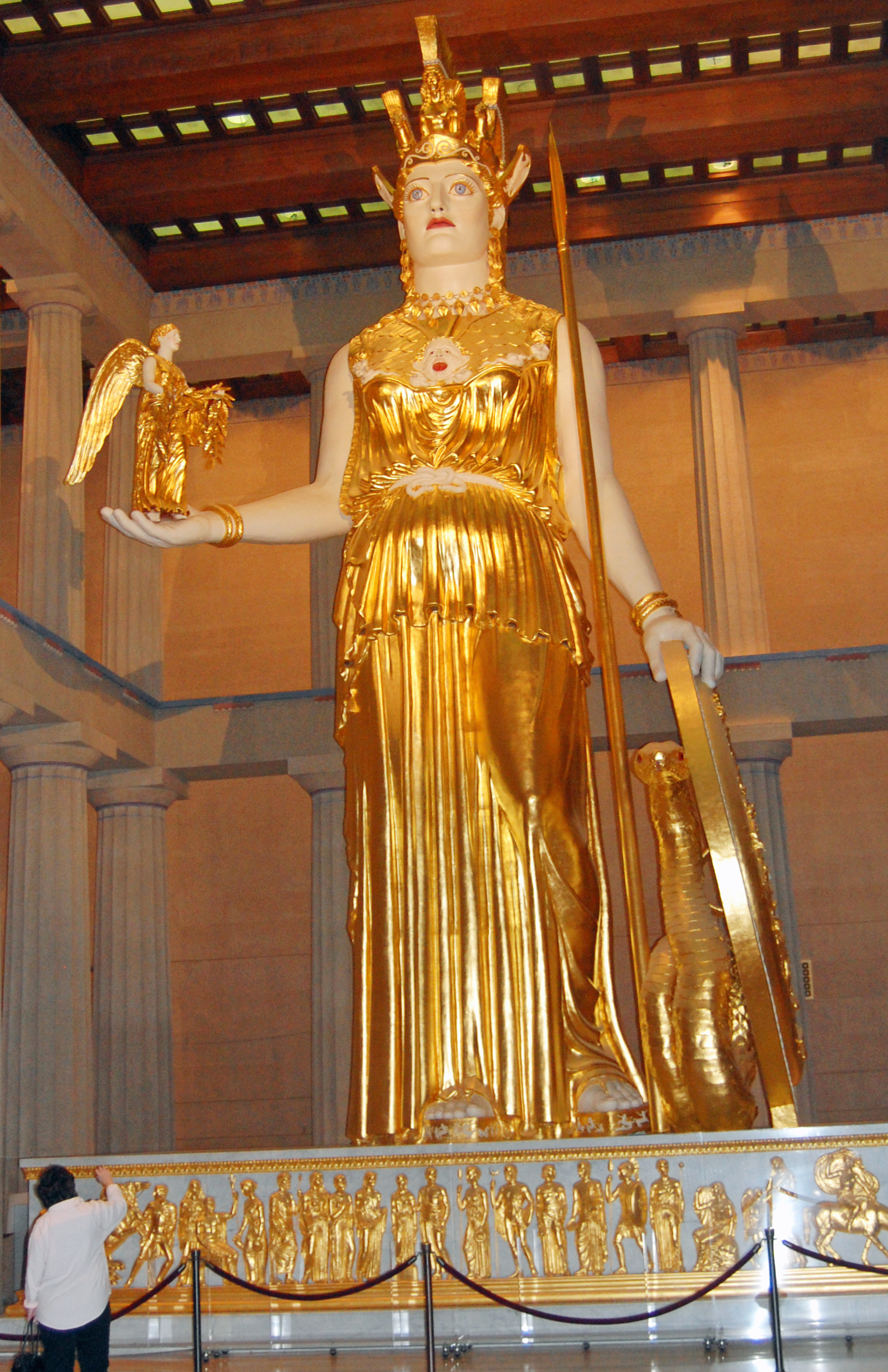 the finished Athena statue in the middle of the Nashville Parthenon. She's very golden, holding Nike in her left hand and a shield in her right hand. She towers above a person standing, looking up at her.