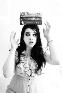 Black and white photo of girl with long dark hair and heavy eye makeup balancing three books on her head, worried expression on face