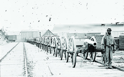 Une photographie montre un soldat afro-américain debout devant une longue file de canons le long d'une voie ferrée.