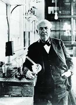 Une photographie montre Thomas Edison dans un atelier bien éclairé. À côté de lui se trouve une table contenant une ampoule à incandescence.
