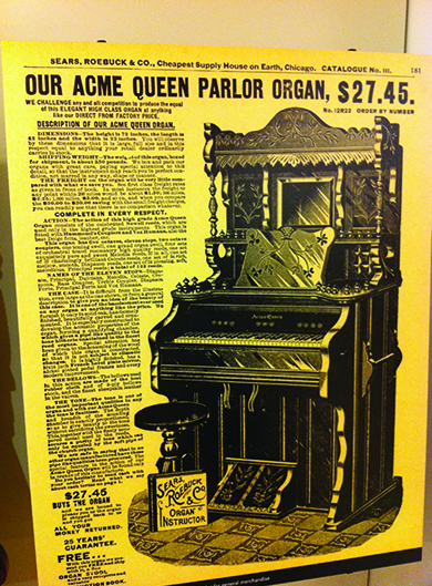 Uma página do catálogo da Sears, Roebuck & Co. anuncia: “Nosso órgão Acme Queen Parlor, $27,45”, seguido por um desenho e uma descrição do produto. O cabeçalho da página diz “Sears, Roebuck & Co., Casa de suprimentos mais barata da Terra, Chicago”.