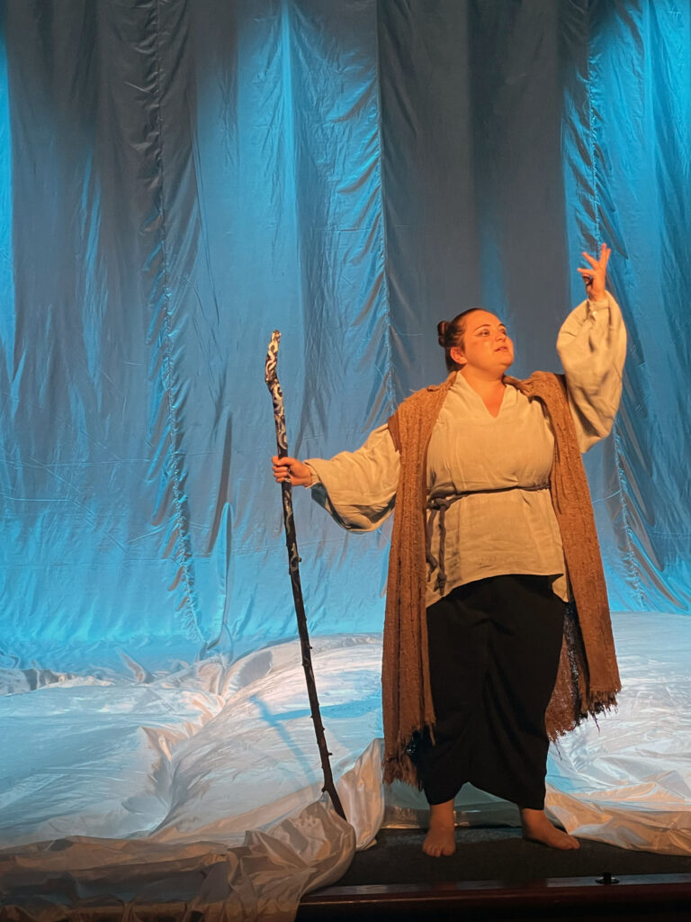 An actor stands on stage in front of a light blue backdrop. They hold a wooden staff in one hand, the end of it touching the ground, and have the other arm extended upwards in a dramatic gesture.