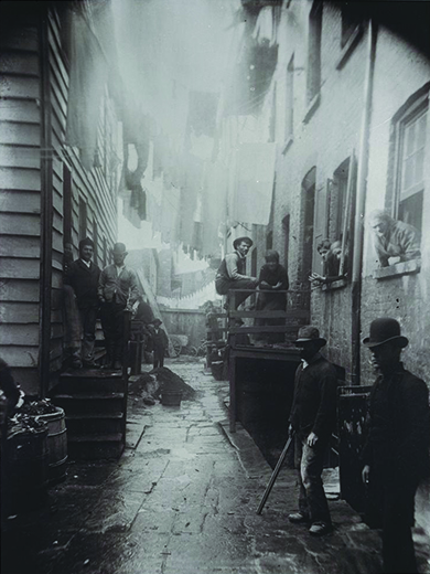 Une photographie montre une ruelle entre deux immeubles. Des hommes, des femmes et des enfants se tiennent debout de chaque côté de l'allée, dans les tabourets et aux fenêtres.