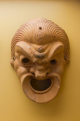 A terracotta comedy mask sculpture. The mask shows a character with a heavily furrowed brow and an open mouth.