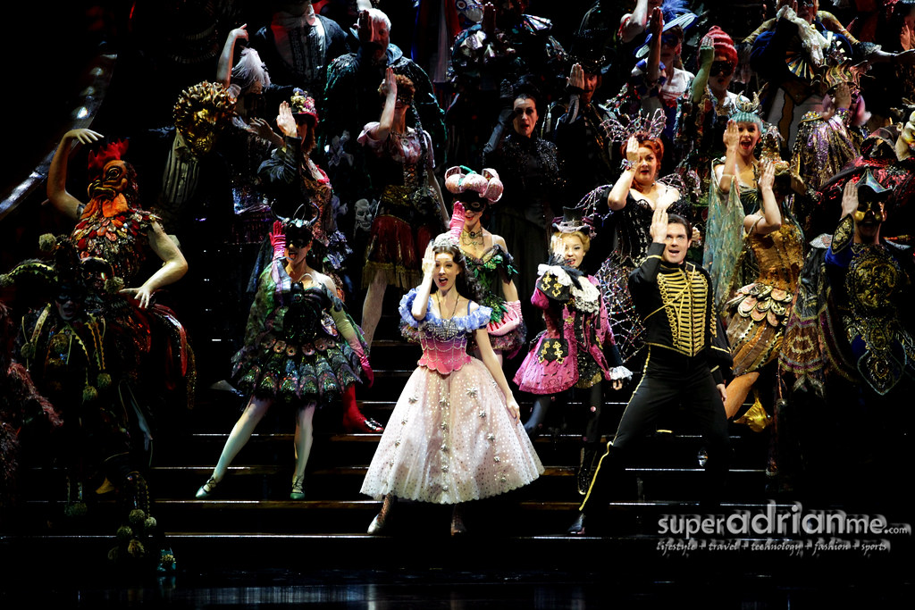 A large group of actors stand on a wide staircase that spans across the entire image during a masquerade ball scene. They wear extravagant costumes and many wear masks. They all are posed in the same manner, with one hand covering one side of their face.