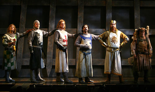 Six men stand in a straight line across a stage. They wear Medieval knight costumes and have their left arm resting on the shoulder of the person standing next to them. The man on the far right end carries a heavy pack.