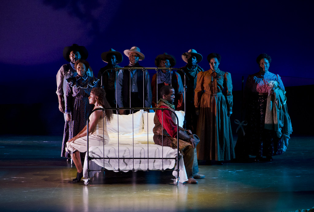 In the center of the image, there is a bed, and two actors sit on the edge of either side facing away from one another. On the left is a woman wearing a white dress, and on the right is a man in a red shirt and brown vest. Behind them stands a group of actors who are dimly lit in a blue light.