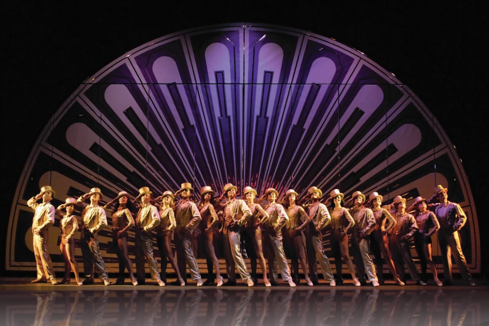 Production photo from "A Chorus Line." On the stage the actors stand in a long straight line all wearing gold costumes with gold top hats. They strike the same pose with one leg extended out, one hand placed on their hip, and the other touching the brim of the hat.