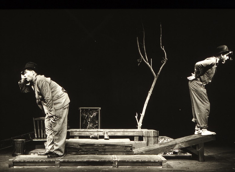 Production photo from Waiting for Godot. The two actors stand at opposite sides of the image, facing away from one another. They are bent over slightly and have a hand shielding their eyes as they look in the distance. In between them is a wooden bench and a small bare tree.