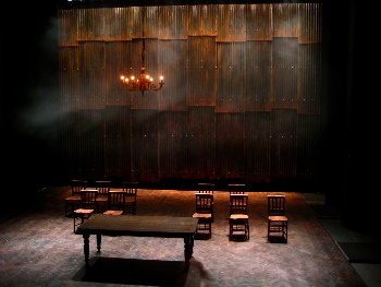 Stage with a long wooden table and six chairs in a line behind it. The lighting is dim and ominous. A simple lit chandelier hangs above the table.