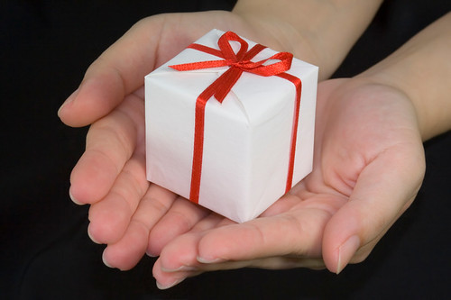 Photo of hands holding out a small white box wrapped with red ribbon