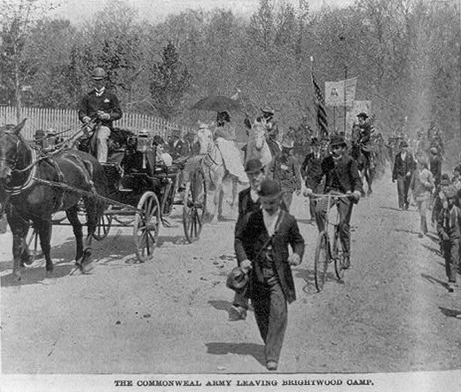 Uma fotografia mostra o Exército de Coxey em marcha, com manifestantes caminhando, montados a cavalo e andando em bicicletas e carrinhos puxados por cavalos.