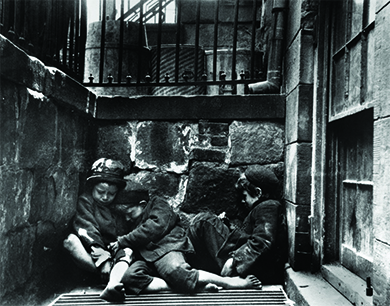 Uma fotografia mostra três crianças pequenas, mal vestidas e descalças, dormindo em uma pilha sobre uma grade de vapor um pouco abaixo do nível da rua.