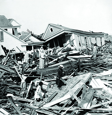 Uma fotografia mostra a devastação do furacão de 1900 em Galveston, Texas. Moradores sobem entre as enormes pilhas de lenha de casas caídas. Várias outras casas destruídas são visíveis ao fundo.