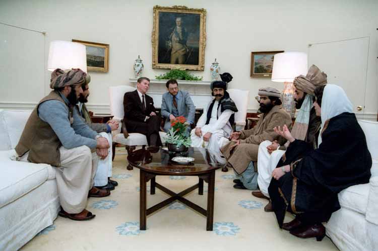US President Reagan met with Afghan Mujahideen leaders in the White House. President Reagan is dressed in a suit. An Afghan woman wearing a head scarf and five male Afghan leaders wearing the traditional peraahan tunbaan and turbans and seated on either side of the US president. Details in text.