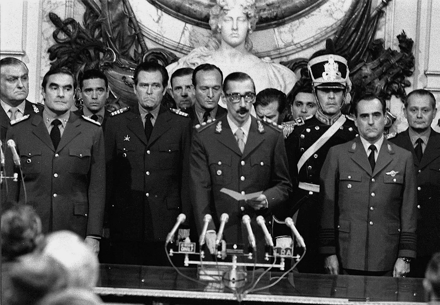 Gen. Videla and his officers dressed in military uniforms. Gen Videla stands in front of microphones for the presidential oath. Details in text.