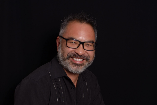 Alejandro has a beard, wears glasses, and is wearing a black shirt.