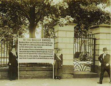 Uma fotografia mostra duas sufragistas em frente ao portão da Casa Branca, segurando uma grande placa entre elas. O texto da placa diz o seguinte: “O presidente Wilson e o enviado Root estão enganando a Rússia. Eles dizem: “Somos uma democracia. Ajude-nos a vencer uma guerra mundial para que as democracias possam sobreviver”. Nós, as mulheres da América, dizemos que a América não é uma democracia. Vinte milhões de mulheres americanas têm o direito de votar negado. O presidente Wilson é o principal oponente de sua emancipação nacional. Ajude-nos a tornar esta nação realmente livre. Diga ao nosso governo que ele deve libertar seu povo antes que possa reivindicar a Rússia como aliada.”