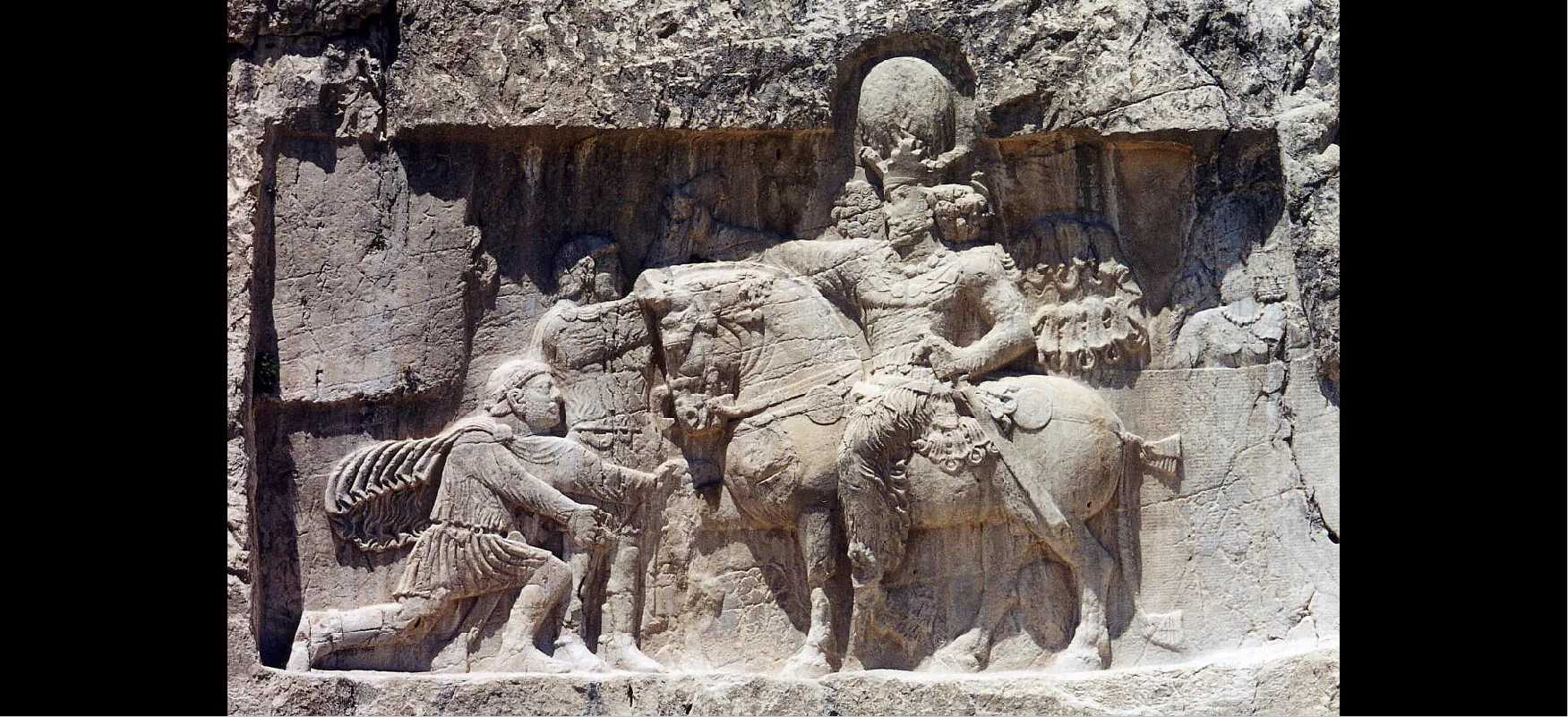 A picture of a wall with a rectangular, beige colored stone carving scene is shown. In the middle, a man is sitting on a horse which is wearing a head dressing with flowers and straps, a decorated saddle, and a bow in its tail. The man is carved with ornate designs all over his torso and legs, has a crown on his head, a beard, flowing curly hair, and a sword on his left side with his left hand on it. His right arm extends over the head of the horse and he holds an object in his hand. Both the man and the horse are looking down at a man kneeling on the ground in front of the horse. He is wearing a flowing cape and a dress of armor. He has a beard and is holding his arms out in front of him. In between the man and the horse stands a man in armor with a beard, his head in the shadows of the carving. The top of a person is carved into the wall behind the horse. Around the rectangular carving is a bumpy stone wall.