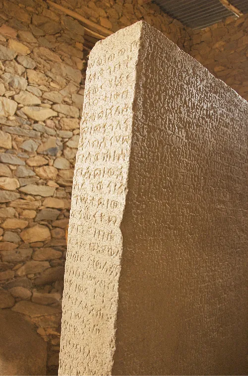 A picture of two sides of a tall, rectangular stone slab is shown against a rocky wall. The stone slab is beige in color and shiny. Words are engraved on both sides of the stone shown. A vent is shown on the ceiling above the stone slab.