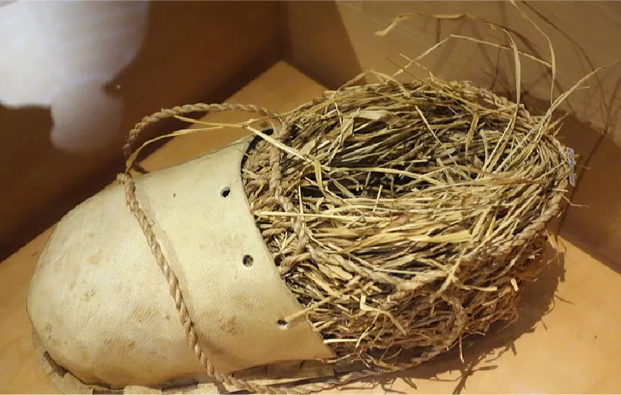 A picture is shown of a circular portion of straw inserted into a beige leather round opening that looks like a shoe. Running from around the straw through holes in the leather is a rope. Rope is also shown laying across the leather portion and secured in the straw section. Under both the straw and the leather, is a piece of leather sewn with brown string. The object is laying on a shiny wood table with brown walls in the background.