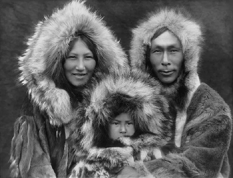 A black and white picture shows three people on a dark background. The woman on the left has small, thin eyes, high cheekbones, and is smiling showing her teeth. The man on the right has almond shaped eyes, high cheekbones, and is shown grinning. The child between them has dark, almond shaped eyes. All three wear thick, furry coats with fur all around the hoods.