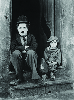 Charlie Chaplin est représenté assis dans l'embrasure d'une porte, les bras croisés, accompagné d'un petit enfant mal habillé.