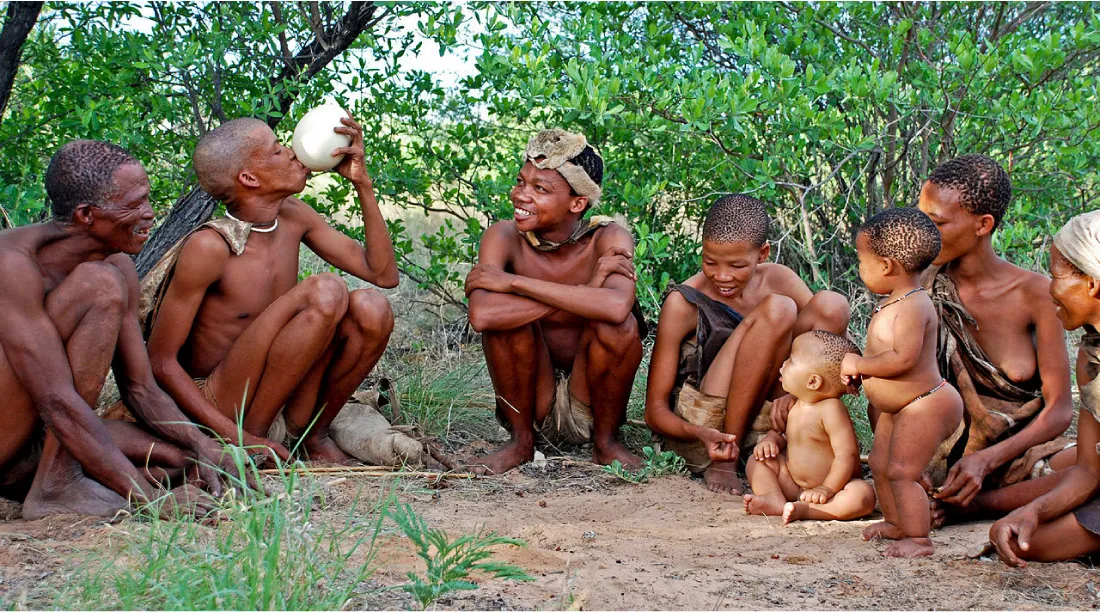 A photo of eight dark skinned people (three men, three women, and two small children) of varying ages are shown siting and squatting on sandy ground in a line amid green leafed trees and grass. The men are naked from the waist up and wear loincloths, the two babies wear beads around their waists while the women wear cloths on one shoulder. Two people wear cloths on their heads. One man drinks form a white jug while everyone else looks and smiles at him.