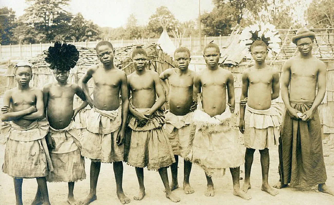 A sepia-colored picture of eight unsmiling people are shown standing in a line on dry ground. They are barefoot, dark skinned, and bare chested with short hair. They all wear rough cloth tied around their waist that goes down to their knees. Some of the cloths have frayed edges and some have ruffles. The last man wears a dark cloth that hangs down to his ankles. The first person wears a square cloth on their head, the second person wears a very dark feathery round object at the front of their head. The seventh person wears a round spray of dark and light feathers on the back of their head and the last man wears a dark bowler hat. In the background there are straw huts, a wooden fence, and trees.