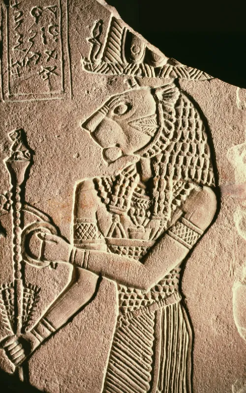 A picture of a carving on a brown, broken piece of stone is shown on a black background. A man is carved with a lion’s head. He wears an elaborately etched headdress, decorated chest armor, and a striped cloth at the waist. Gridded and plain bands adorn his arms and wrists and he holds a twisted scepter with a small seated lion on top in his right hand. Above his head are etchings and ancient writings in a rectangular box.