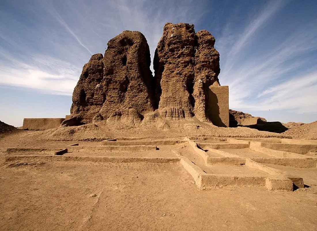 A picture of a building made of sandstone is shown. Four tall, dark brown, rough and bumpy projections stand up from the ground with some rectangular walls at both sides. In the forefront lays a maze of short, sandy walls low to the sandy ground. Blue skies with long thin clouds show in the background.