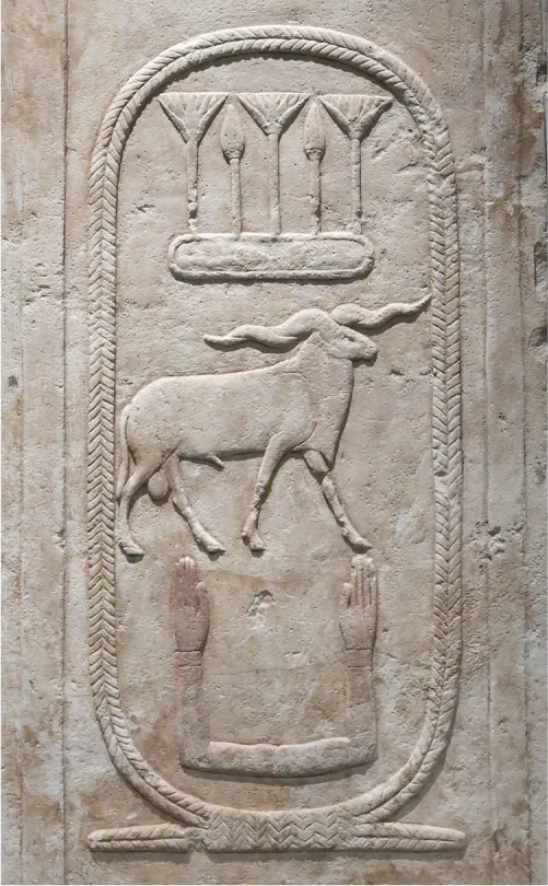 A broken piece of a long rectangular stone shown vertical on a solid black background is pictured. Hieroglyphs are carved from the top to the bottom. There is an oval with a pedestal at the bottom and three carvings inside. At the top is a decorative carving that has three tall broom-like projections and then two smaller spears in between them, all placed on a horizontal oval etching. Next is the carving of a large four-legged animal resembling a ram with long twisted horns on the sides of his head. At the bottom are two arms connected to each other in a “U” shape with palms facing straight up.