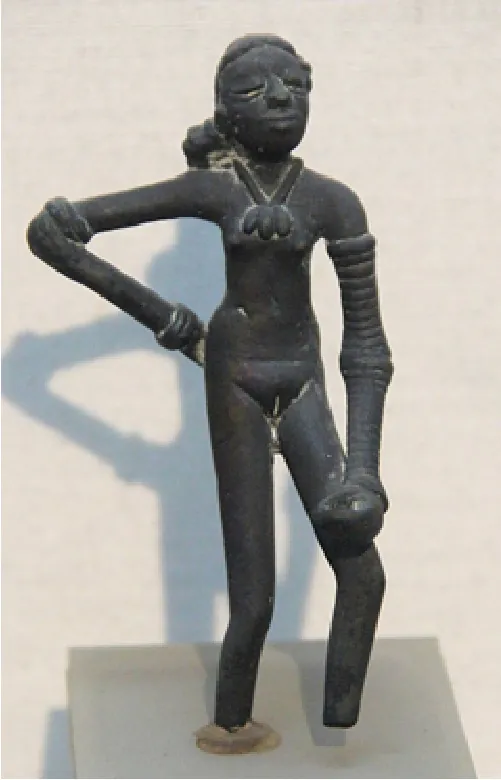 An image of a dark gray figurine is shown on a gray square pedestal and off white background. The figurine is thin with long arms and legs. She shows closed, swollen eyes, long hair in a side bun, and a half smile. A necklace hangs around her neck with three leaf shaped projections hanging above her breasts. She is naked and has bracelets on her right wrist and above her right elbow and her left arm shows bracelets from wrist to armpit. Her left hand is large and closed in a fist, resting on her left knee. Both her feet are missing and her left leg is hanging in the air while the right leg is anchored to a small, brown, round mound.
