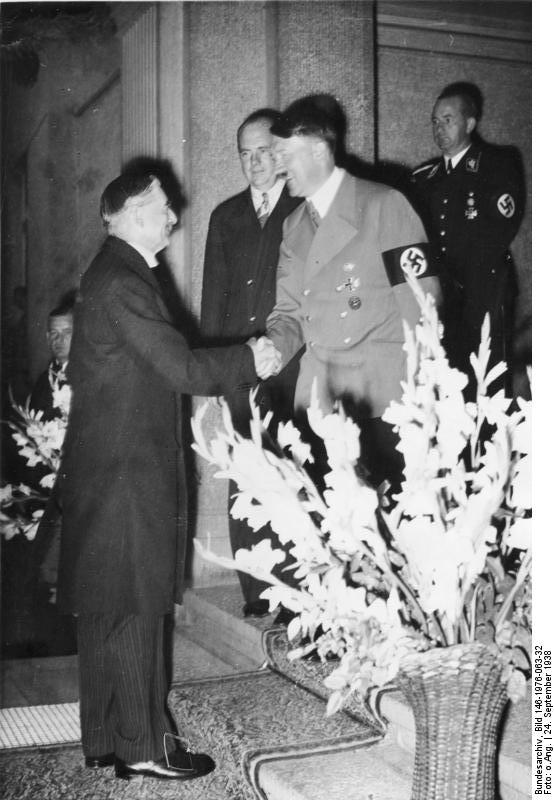 Prime Minister Chamberlain smiling and shaking hands with an equally cheerful Adolf Hitler.