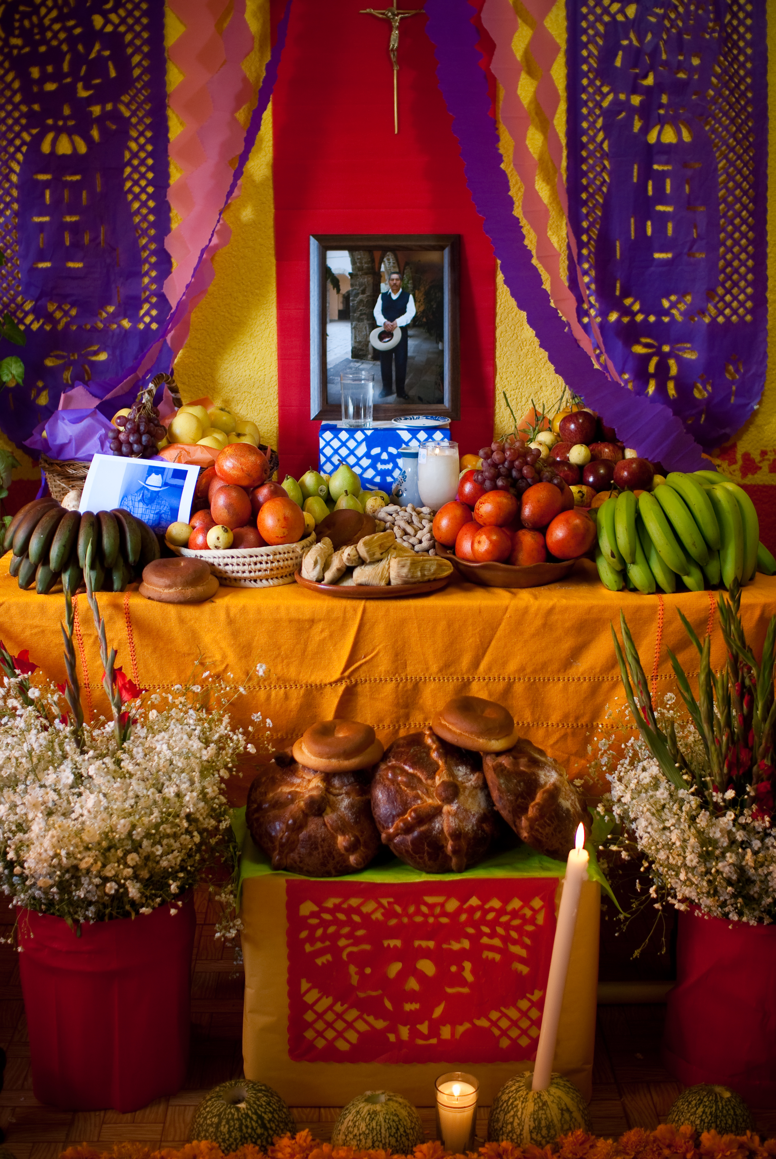 Table covered with an orange tablecloth and various foods. There is also a photo of an older man in traditional Mexican dress and purple paper cuttings hung from the ceiling.