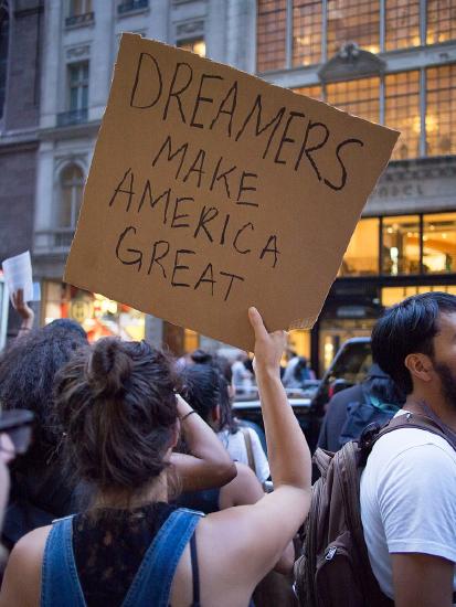 A cardboard sign with the handwritten message "Dreamers make America great" held up by a woman in a crowd outside a multi-story lighted building.