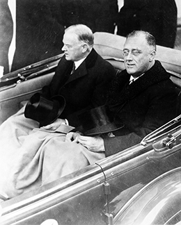 Une photographie montre Herbert Hoover et Franklin D. Roosevelt roulant côte à côte à l'arrière d'un véhicule décapotable. Une couverture couvre leurs jambes et leurs chapeaux reposent sur leurs genoux.