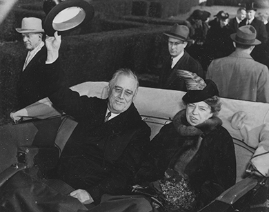 Uma fotografia mostra Franklin e Eleanor Roosevelt sorrindo enquanto andam na traseira de um ônibus. Franklin Roosevelt acena com seu chapéu para os espectadores.