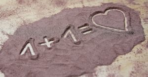 Picture of sand on the ground and someone wrote 1 + 1 = heart in it.