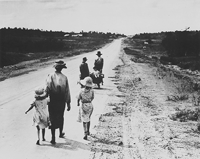 Una fotografía muestra a seis refugiados de Dust Bowl, tres adultos, dos niños y un bebé, caminando por una carretera. El bebé cabalga en una carreta pequeña.