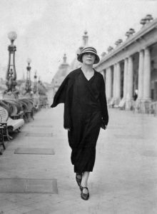 Una mujer vestida de negro, con sombrero camina hacia la cámara en un paisaje urbano.