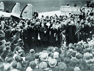 Una fotografía muestra a Neville Chamberlain inmediatamente después de su llegada a Inglaterra, donde se dirige a una entusiasta multitud de funcionarios y prensa.