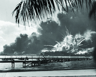Une photographie montre un long quai avec l'USS Shaw qui explose derrière lui. Au fond, d'énormes vagues de fumée sont visibles.