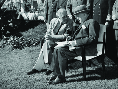 Une photographie montre Winston Churchill et le président Roosevelt assis à l'extérieur sur des chaises, réunis autour de journaux, avec une rangée de fonctionnaires debout derrière eux.