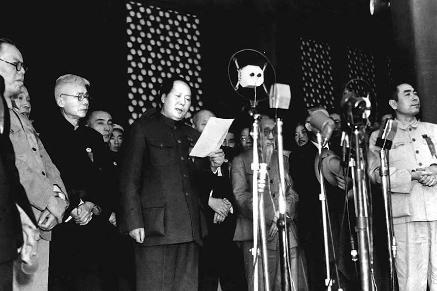 A photograph shows men standing behind several standing microphones. The men are of Asian descent, wearing suits. Some have glasses on. They are standing listening to one man in the middle of the picture read from a white piece of paper into the microphones.