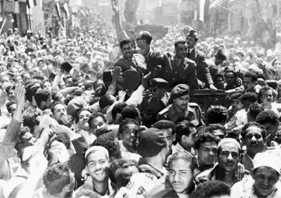 A photograph shows a crowd, mostly of men. In the middle of the crowd, seven men are riding in a car looking at the crowd. They wear suits and hats. People from the crowd are reaching out to touch them. Men in military gear and caps walk alongside the car looking down and at the crowd.