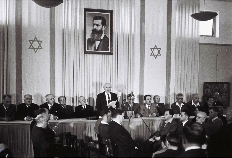 A photograph shows several men sitting behind a long table. A man in the middle stands, holds a piece of paper, and speaks into a microphone. In front of the table, groups of men sit in chairs. On the wall behind the table are long drapes, banners with a six pointed star, and a large portrait of a man in a suit, bow tie, and beard.
