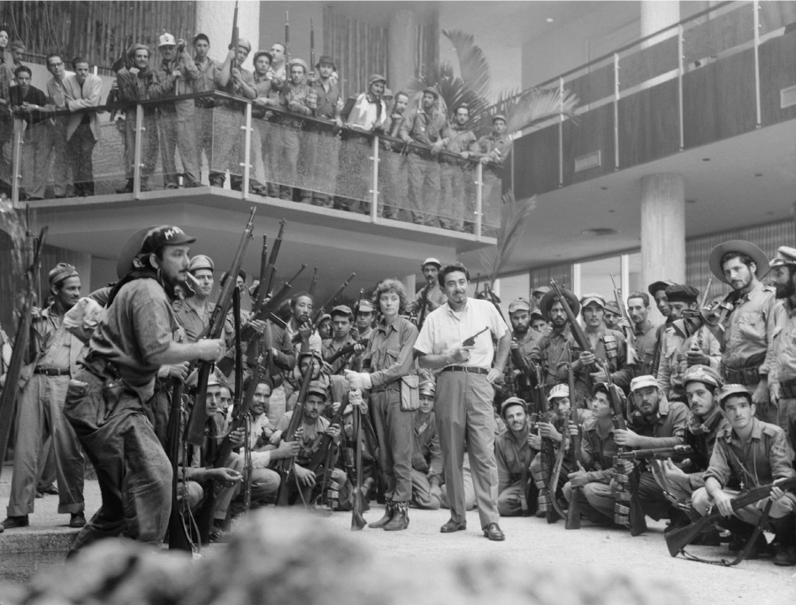 A photograph shows a man standing inside a building in a white short sleeved shirt and pants, holding a pistol in his right hand and smiling. Behind him stands a woman in pants holding a rifle while it rests on the floor. Surrounding them in several rows in a semi-circle is a large group of men, all holding rifles or other weapons. Above them on a platform with a glass railing are more men with weapons looking down at the group below.